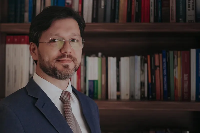 Retrato de advogado do escritorio, de terno, diante de uma estante de livros
