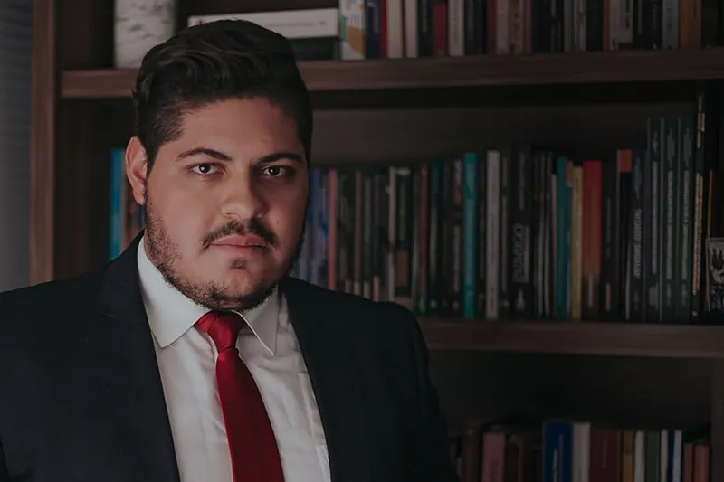 Retrato de advogado do escritorio, de terno e gravata, diante de uma estante de livros